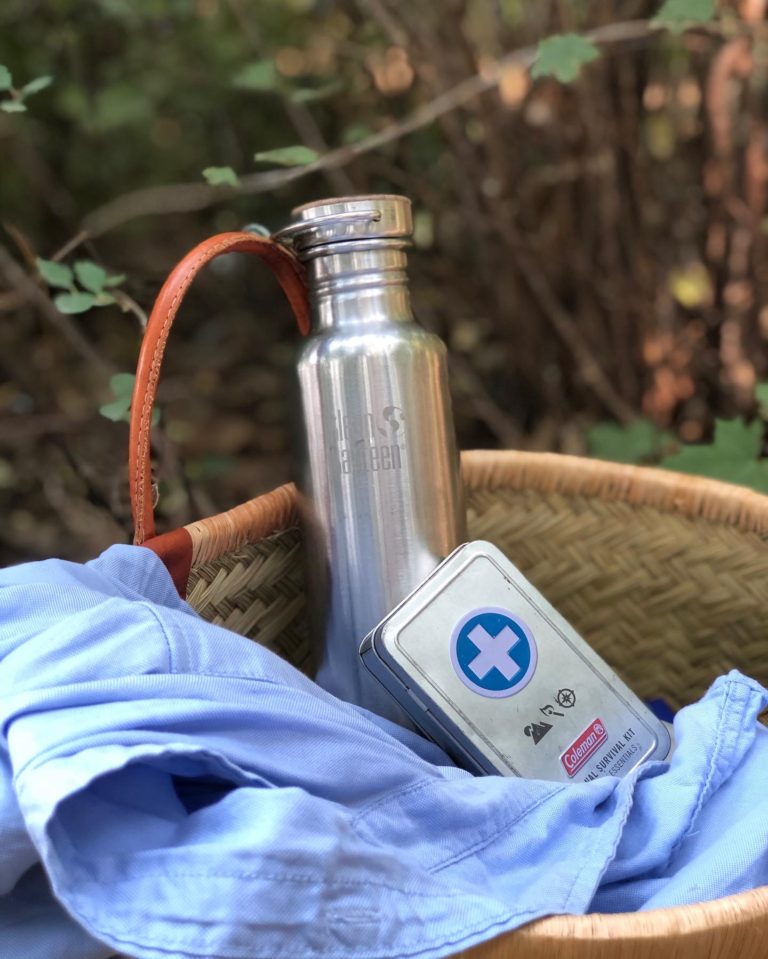 Klean-Kanteen stainless steel water bottle, Coleman-USA-first-aid kit, j crew blue Oxford shirt and picnic basket next to a lake